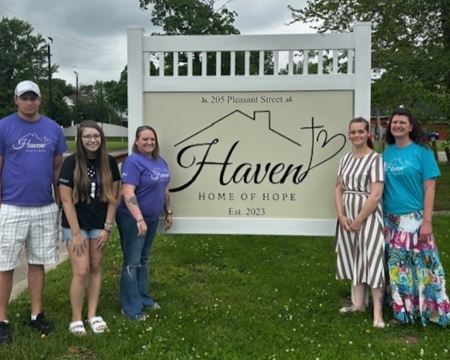 five persons involved with supporting and staffing Haven Home stand beside the house's large business sign in the green lawn.