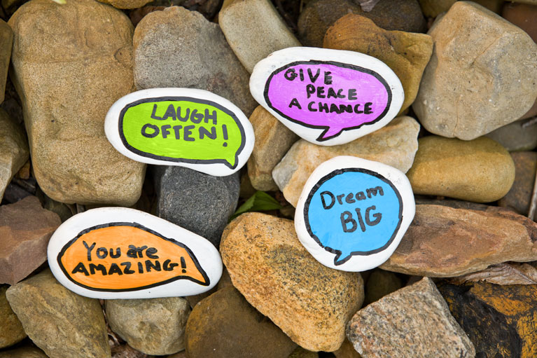 brown stones beneath several stones painted white with colorful speech bubbles with inspiring positive phrases.