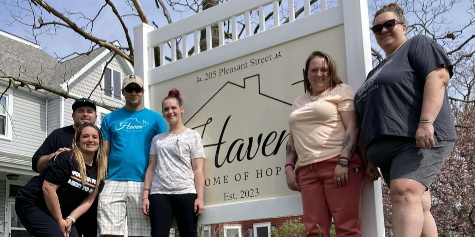 men and women stand smiling outdoors in residential neighborhood in front of large yard sign that reads Haven Home of Hope.