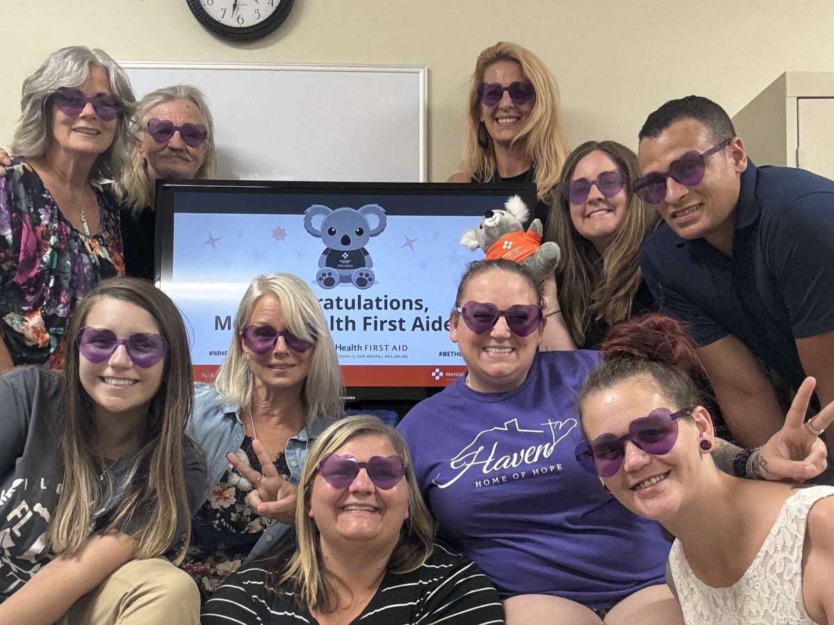 9 women and 1 man pose in a small office classroom wearing sunglasses with purple heart frames