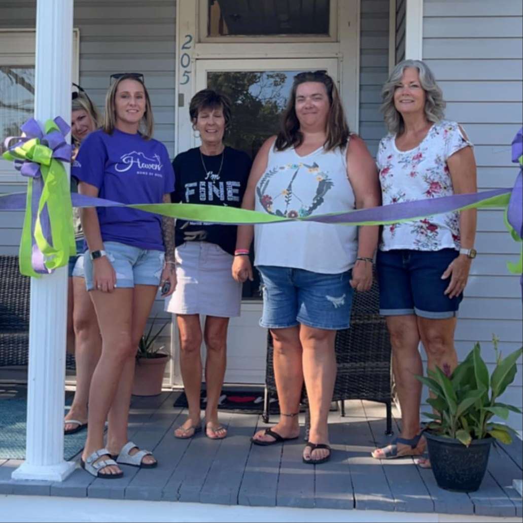 five women in tee shirts, shorts and sandals on a residential porch standing behind a large green and purple horizontal ribbon
