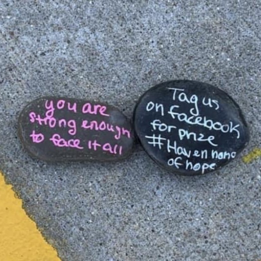 two flat smooth stones placed on a parking lot surface next to a yellow line, bright pink and green words are written in marker on stone faces